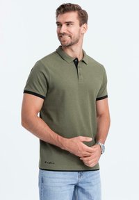 Zelené polo tričko s černými akcenty, krátké rukávy, tříknoflíková léga a detail loga na lemu. Nosené s světle modrými džíny.