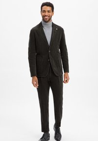 Dunkelbrauner Cordanzug mit taillierter Jacke, Ein-Knopf-Verschluss und passenden Hosen. Getragen mit einem hellgrauen Rollkragenpullover und schwarzen Schuhen.