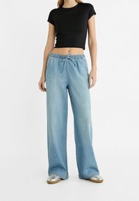 Ljust blå denimbyxor med vida ben, dragsko i midjan och sidofickor, kombinerade med en svart åtsittande crop top och metalliska skor.