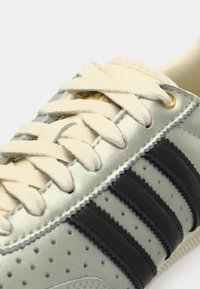 Srebrny sneaker z czarnymi paskami, skórzana cholewka, perforowane detale, kremowe sznurówki i złote akcenty przy oczkach.