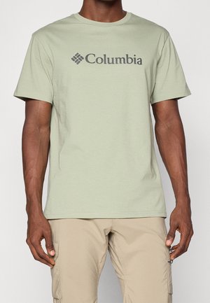 Hombre con camiseta Columbia verde claro y pantalones cargo beige, de pie frente a un fondo liso con los brazos relajados a los lados.