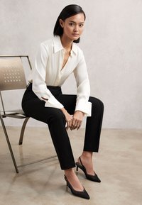 Ung kvinde med sort hår sidder på metalstol iført hvid bluse, sorte bukser og sorte slingback-højhælede sko mod en ensfarvet væg.