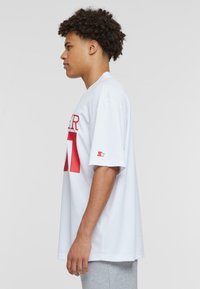 Camiseta blanca de manga corta con letras y número en rojo. Corte holgado, cuello redondo, con un logo destacado en la manga izquierda. Material suave de jersey.