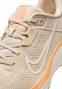 Zapatilla deportiva de color beige claro con una parte superior de malla texturizada, acentos blancos translúcidos y un logo naranja en la lengüeta. Detalles perforados notables.