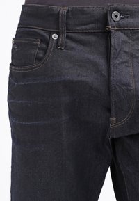 Jeans de mezclilla oscura con una textura suave, cinco bolsillos y costuras naranjas en contraste. Cierre frontal con botón y cremallera.