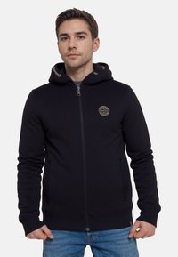 Hombre joven vestido con una sudadera con cremallera negra y un logo circular beige en el pecho, y jeans de color azul claro, de pie contra un fondo blanco.