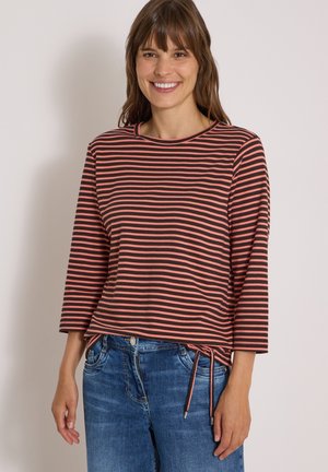 Femme souriante portant un top à manches longues rayé rose et noir avec des liens latéraux et un jean denim bleu sur un fond clair uni.