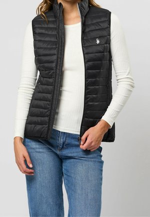 Vrouw draagt een opengezette zwarte doorgestikte bodywarmer over een wit geribbeld shirt met lange mouwen en een blauwe spijkerbroek, houdt de rits van de bodywarmer met één hand vast.