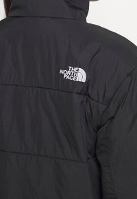 Black puffer jacket with a high collar, featuring a white embroidered logo on the left shoulder and a smooth, textured fabric.