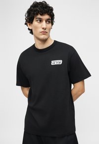 Άνδρας με σγουρά σκούρα μαλλιά που φοράει μαύρο t-shirt Versace Jeans Couture, κοιτάζοντας προς τα πλάγια με τα χέρια πίσω από την πλάτη του.