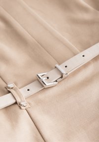 Tissu beige avec une ceinture en cuir blanc dotée d'une boucle argentée, texture plate, et clous en métal pour la fixation. Design simple, sans motifs.