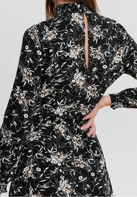 Schwarzes Blumenkleid mit langen Ärmeln, rüschigem Ausschnitt und offenem Rückendetail. Verziert mit weißen und orangefarbenen Blumenmustern auf weichem Stoff.