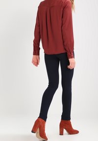 Mörkröd långärmad blus med axelkotttar, kombinerad med marinblå byxor och oranga mocka klackkängor med blockklack. Slät textur, figurnära design.