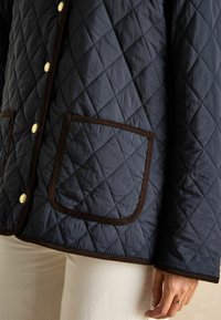 Granatowa pikowana kurtka w diamentowy wzór, z brązowym wykończeniem i złotymi zatrzaskami; posiada dwie kwadratowe kieszenie. Teksturowany materiał.