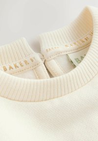 Krémový sveter s rebrovaným golierom, na ktorý je na výstrihu zlatým embosovaným písmom napísané "BAKER". Mäkká textúra a jednoduchý dizajn.