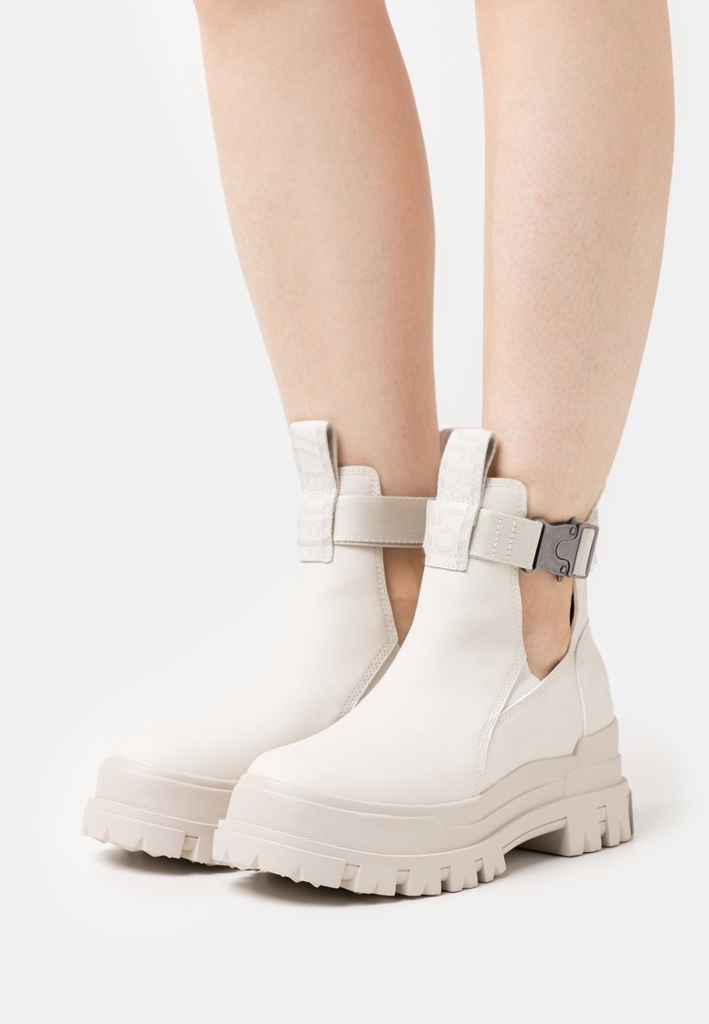 Bottines blanches à cheville avec panneaux latéraux élastiques, semelle épaisse texturée, et une bride avec un détail de boucle métallique. Finition en matériau lisse.