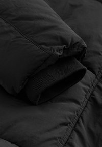 Jachetă puffer, izolată, de culoare neagră, cu o carcasă exterioară texturată și manșete negre ribbate, având un finisaj neted și detalii cu cusături.