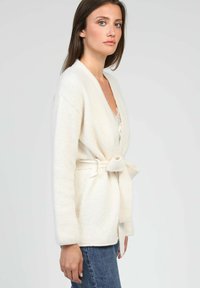 Cardigan en tricot beige avec une ceinture à nouer, décolleté en V, manches longues et texture douce. Porté sur un haut clair, associé à un jean bleu.