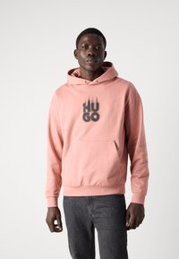 Sudadera rosa con un bolsillo frontal y capucha con cordón. Texto negro "HUGO" impreso en una fuente gruesa y estilizada. Tejido suave y corte relajado.