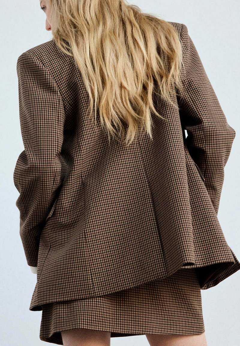 Femme blonde en blazer marron à carreaux et mini-jupe assortie, tournée de dos devant un fond uni.