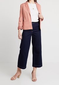 Vaaleanpunainen bleiseri, harmaa t-paita, tummansiniset leveät housut ja nude-sandaalit. Bleiserissä on yksi nappi ja käärityt hihat.