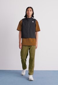 Man walking wearing black zip-up vest, brown short-sleeve shirt, olive green pants, and white sneakers against plain background.