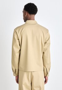 Chaqueta de algodón ligera en color beige, con cuello, mangas largas, puños abotonados y un corte relajado, mostrada desde la parte de atrás.