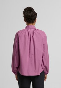 Langärmlige Bluse mit hohem Kragen aus hellrosa Stoff. Verfügt über einen gerafften Rückeneinsatz und elastische Bündchen, mit einer lockeren Passform.