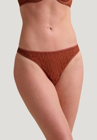 Tanga en tissu marron texturé avec un motif géométrique. Il dispose d'une taille lisse et de fines lanières sur les côtés pour une couverture minimale.