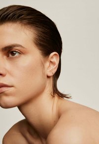 Pendientes de oro con una cadena y un colgante puntiagudo. El modelo tiene el pelo corto y húmedo; la piel tiene una textura natural y tonos suaves.