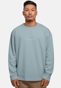 Felpa azzurro chiaro con scollatura a girocollo, maniche lunghe e un logo sottile in rosa. Tessuto morbido con una consistenza liscia.