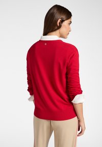 Nainen, jolla on yllään punainen neule valkoisen paidan päällä, hihat käärittyinä ja vaaleat housut, kääntyy poispäin yksinkertaista taustaa vasten.