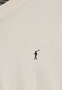 Sweter pulpowy na drutach w kolorze ecru z dekoltem w serek, z małą czarną haftowaną postacią trzymającą kij golfowy na piersi.