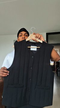 Sort ærmeløs vest med vertikal quiltet design, to frontlommer og sorte knapper. Hænger fra en træstang.