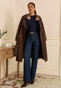 Hnědý suedový trenčkot, námořnický žebrovaný svetr s vlnitým výstřihem, tmavé džíny s širokými nohavicemi a hnědé lodičky se špičatou špičkou, stojící uvnitř.