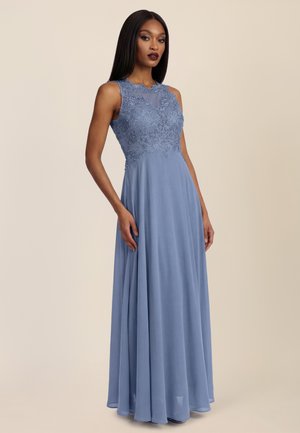 Frau in ärmellosem staubblauem Kleid mit floraler Spitzenoberfläche und fließendem Chiffonrock, stehend vor einfachem beigem Hintergrund.