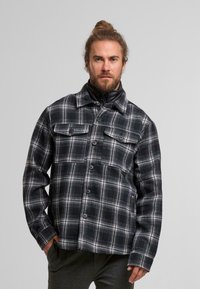 Karierte Flanelljacke in Schwarz und Weiß mit grauen Akzenten, ausgestattet mit einem Kragen, Knopfverschluss und zwei Brusttaschen. Strukturierter Stoff.