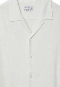 Camicia bianca in lino con vestibilità comoda, presenta un colletto, una fila di bottoni e una texture morbida; l'etichetta indica la taglia L.