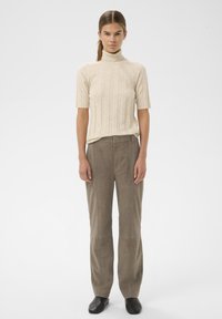 Vrouw staand, gekleed in een beige coltrui met korte mouwen, taupekleurige hoge taille broek en zwarte instapschoenen, op een witte achtergrond.