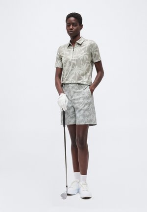 Isik, kes kannab ühtse palmi- mustrilise golfisärgi ja -lühikeste pükstega, hoides golfikeppi kinnastega käes, seisab ühevärvilise tausta ees.