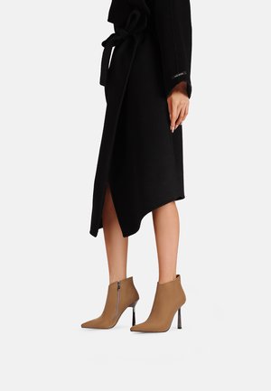 Stivaletti alti beige con tacco a spillo e punte affusolate, con cerniere laterali e tacchi metallici, abbinati a un cappotto avvolgente nero.