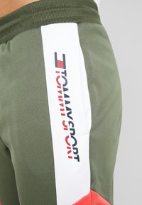 Närbild av olivgröna sportbyxor med vit vertikal rand som visar "Tommy Sport"-logotypen i marinblå och röd text.