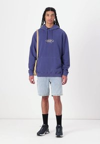 Fialová mikina s predným logom, skombinovaná s svetlými denimovými šortkami. Čierne ponožky a tenisky dopĺňajú outfit. Model nosí béžovú messenger tašku.
