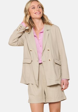 Beige linen blazer with double-breasted design, two front pockets, and button accents. Worn over a light pink shirt and beige shorts.