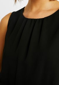 Detailní záběr na osobu s bezrukávovým černým topem s nařasením v oblasti kulatého výstřihu.