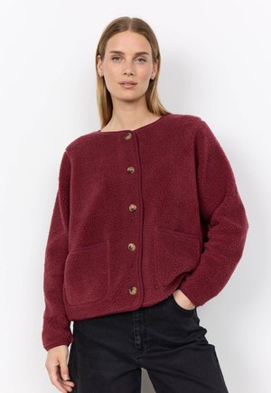 Woman wearing a burgundy button-up fleece jacket with front pockets, posing with one hand in black pants pocket against a white background.