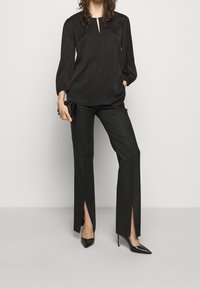 Blouse noire avec un col en trou de serrure et des manches bouffantes, associée à un pantalon noir ajusté avec des fentes sur les côtés. Escarpins noirs à talons pointus inclus.