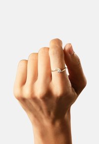 Dünner Goldring mit einem zentralen runden Diamanten, umgeben von einem verdrehten Design auf beiden Seiten. Die Hand präsentiert den Ring vor einem hellen Hintergrund.