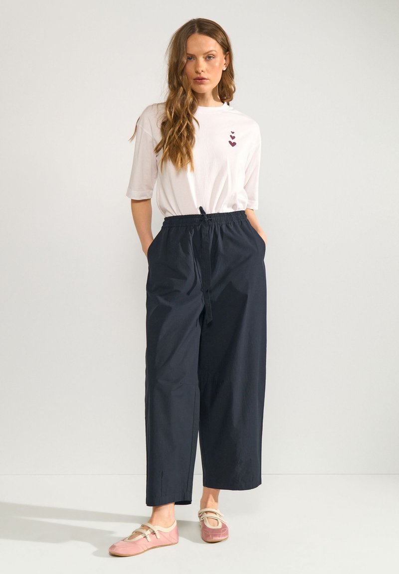 Femme aux longs cheveux ondulés portant un t-shirt blanc avec trois impressions de cœurs, un pantalon noir ample et des chaussures plates roses sur un fond uni.