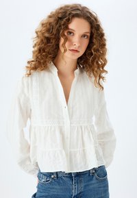Witte katoenen blouse met knoopsluiting aan de voorkant, kantaccenten en een gelaagd ontwerp. Heeft lange mouwen en een ontspannen pasvorm, gecombineerd met een blauwe spijkerbroek.